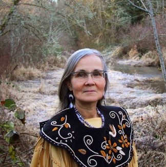A Native woman with grey hair, glasses, and an embroidered shirt stands in front of a stream in the woods.